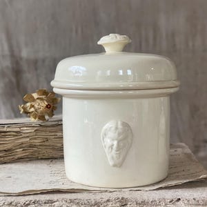 以下が含まれることがあります： クリーム色のセラミックジャー。蓋と前面に装飾的な顔があります。ジャーは丸い形で、小さな花のようなノブが付いた蓋が付いています。ジャーは古い紙の束の上に置かれています。