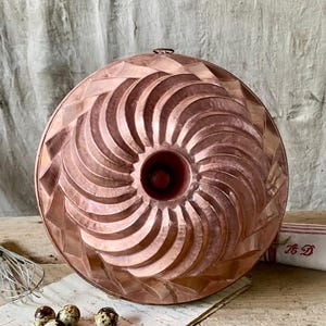 May include: A copper Bundt tin with a spiral design. The tin has a central tube and a decorative pattern on the exterior. The copper has a warm, reddish-brown hue. A whisk and some eggs are in the background.