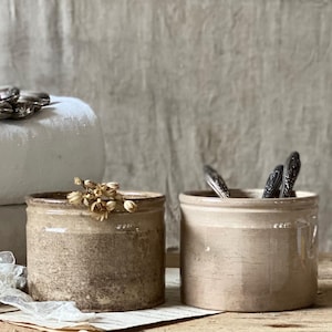May include: Two antique stoneware crocks in shades of beige and brown sit on a wooden surface. One crock has dried flowers, the other has silverware. The crocks have a rustic, aged appearance, suggesting they are vintage.