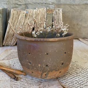 May include: A rustic, brown ceramic pot with small holes, filled with dried white flowers. The pot has a weathered appearance and sits on a surface with aged paper and wooden objects.