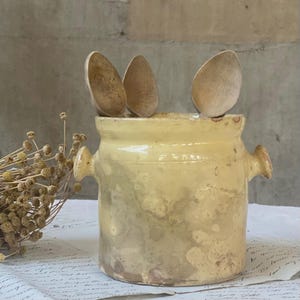 Peut inclure: Un pot en céramique jaune pâle doté de deux anses latérales contenant trois cuillères en bois brun clair. Le pot présente une finition émaillée lisse avec des variations subtiles de couleur et de texture, et les cuillères sont placées verticalement.
