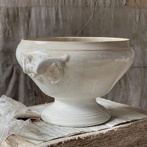 May include: Cream-coloured ceramic serving bowl with a pedestal base and decorative floral handles. The bowl has a wide rim and a slightly flared body. The surface has a subtle, aged appearance, suggesting a vintage or antique aesthetic. The bowl is likely used for serving food.
