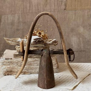May include: A rustic metal bell attached to a curved wooden yoke, resting atop a stack of weathered, aged paper documents. The bell has a dark, oxidized finish, and the wooden frame features a natural, textured grain with visible signs of wear.