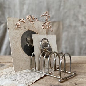 Pode incluir: Um porta-cartas de metal prateado com um design curvo, segurando fotografias vintage e flores secas. As fotografias apresentam um retrato de um homem. O porta-cartas está sobre uma superfície de madeira.