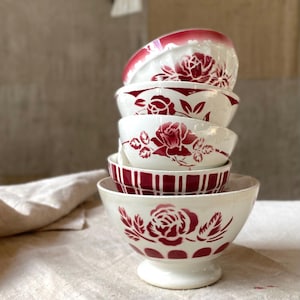 Può includere: Una pila di cinque ciotole in ceramica bianca con motivi di rose rosso scuro e disegni geometrici. Le ciotole variano in dimensioni e sono disposte verticalmente, ognuna con un distinto motivo floreale o a griglia in una combinazione rosso e bianco.