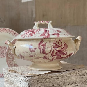 May include: A cream-coloured ceramic tureen featuring a deep red floral transferware pattern. The lidded vessel has two side handles and a matching lid with a decorative handle. A matching plate with a red border pattern is visible behind the tureen.