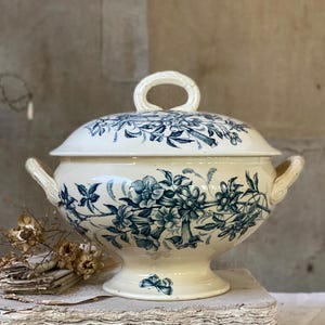 May include: A cream-coloured ceramic tureen with a matching lid, featuring dark blue floral patterns and a small butterfly motif on the base. The vessel has two side handles and a central loop handle on the lid, all with a textured, twisted design.