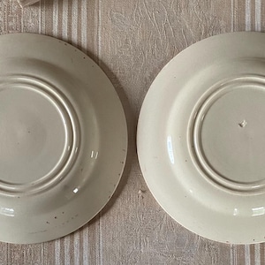 May include: Two off-white ceramic plates, viewed from the underside, are arranged side-by-side. Each plate features a central recessed area surrounded by concentric rings. The plates have a slightly aged appearance with subtle imperfections.