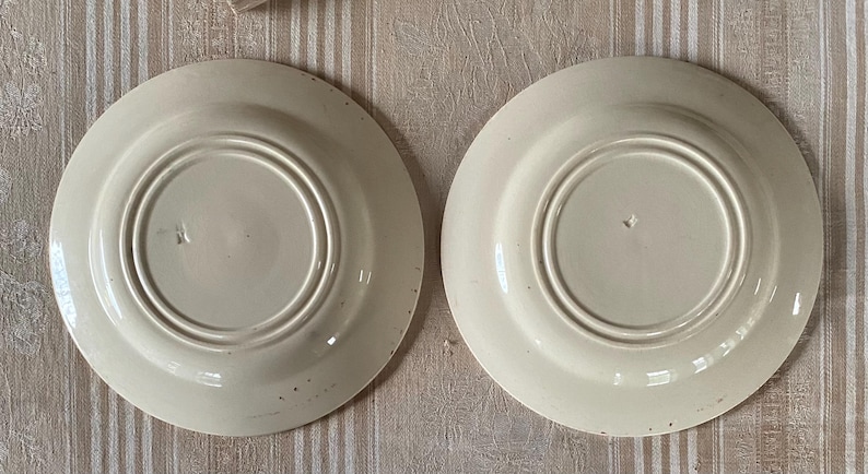 May include: Two off-white ceramic plates, viewed from the underside, are arranged side-by-side. Each plate features a central recessed area surrounded by concentric rings. The plates have a slightly aged appearance with subtle imperfections.