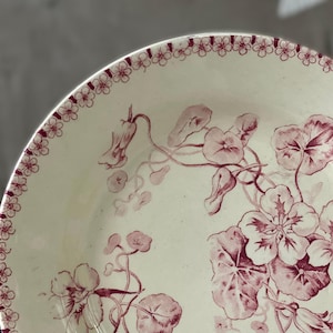 May include: Cream-colored ceramic plate with a floral design in shades of burgundy. The plate features a border of small flowers and a detailed illustration of flowers and leaves. A vintage-style decorative piece.