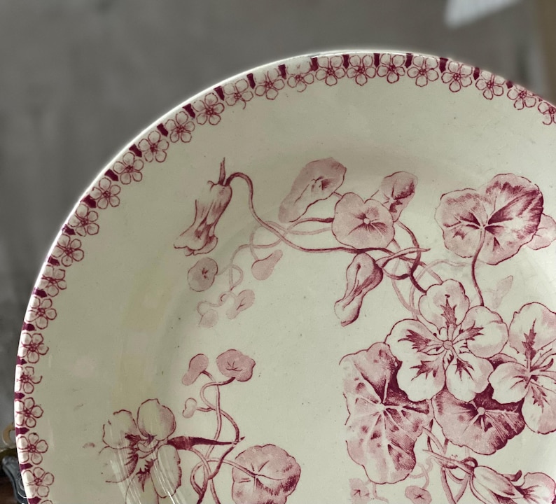 May include: Cream-colored ceramic plate with a floral design in shades of burgundy. The plate features a border of small flowers and a detailed illustration of flowers and leaves. A vintage-style decorative piece.
