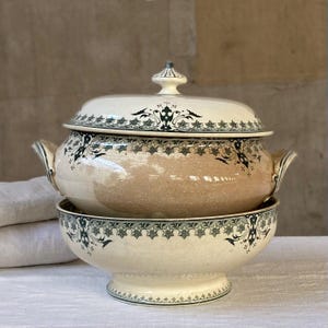 May include: A ceramic tureen with a matching lid, featuring a cream-coloured base with a subtle crackle glaze finish. The piece is decorated with a dark green floral and scrollwork border pattern, two side handles, and a central lid knob.