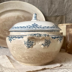 May include: Cream ceramic tureen with a lid, featuring blue floral patterns and a decorative handle. The round tureen sits on a table with vintage paper. The tureen is approximately 25 cm in diameter.