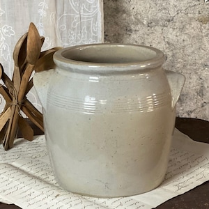 Puede incluir: Una vasija de gres gris claro con cuerpo cilíndrico, dos asas laterales y tres líneas horizontales incisas alrededor del cuello superior. El recipiente de cerámica tiene un acabado liso y moteado con un borde superior ancho y abierto.