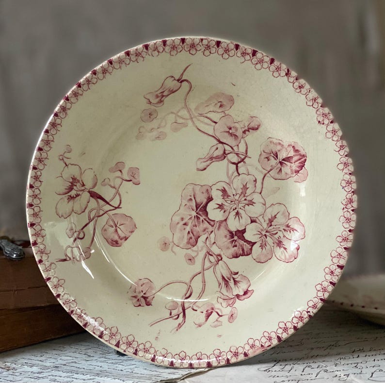 May include: Cream-colored ceramic plate with a floral design in shades of red. The plate features a border of small red flowers and a central pattern of stylized flowers and leaves. A vintage-style decorative piece.