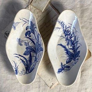 Könnte beinhalten: Zwei antike, birnenförmige Keramikschalen mit blauen Blumenmustern. Die weißen Schalen zeigen detaillierte blaue Illustrationen von Blumen und Blättern. Die Schalen liegen auf gealtertem Papier, was ihre Vintage-Ästhetik unterstreicht.