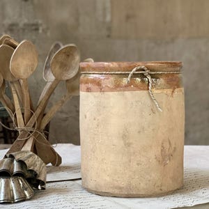 May include: A cylindrical stoneware crock with a beige body and a brown glazed rim, tied with a piece of twine. Beside it sits a bundle of wooden spoons and a stack of metal fluted molds, all resting on a surface covered with vintage paper.