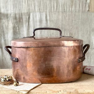 以下が含まれることがあります： 蓋と両側の取っ手が付いた銅製の鍋。鍋の上部には濃い色の取っ手があり、木製の表面に置かれています。鍋は丸く、光沢のある反射面を持っています。