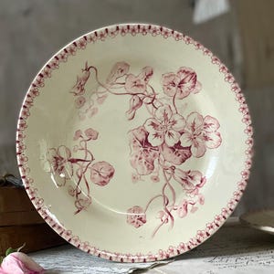 May include: Cream-colored ceramic plate with a floral design in shades of burgundy. The plate features a detailed pattern of flowers and leaves, with a delicate border of small floral motifs. A vintage-style decorative piece.