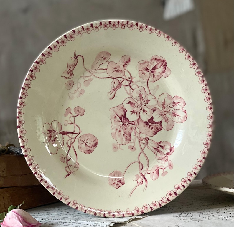 May include: Cream-colored ceramic plate with a floral design in shades of burgundy. The plate features a detailed pattern of flowers and leaves, with a delicate border of small floral motifs. A vintage-style decorative piece.
