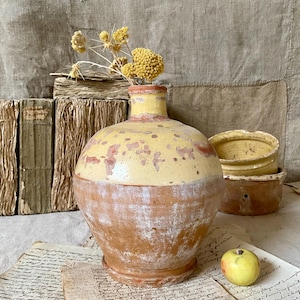 Może przedstawiać: Rustykalny, dwukolorowy ceramiczny wazon z żółtą górą i terakotowym dołem. Wazon zawiera suszone żółte kwiaty. Dodatkowe terakotowe donice i jabłko są widoczne w tle, wraz ze starymi książkami i papierami.