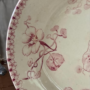 May include: Cream-colored ceramic plate with a floral design in burgundy. The plate features a border of small floral patterns and larger flowers with vines and leaves. A classic, vintage aesthetic.