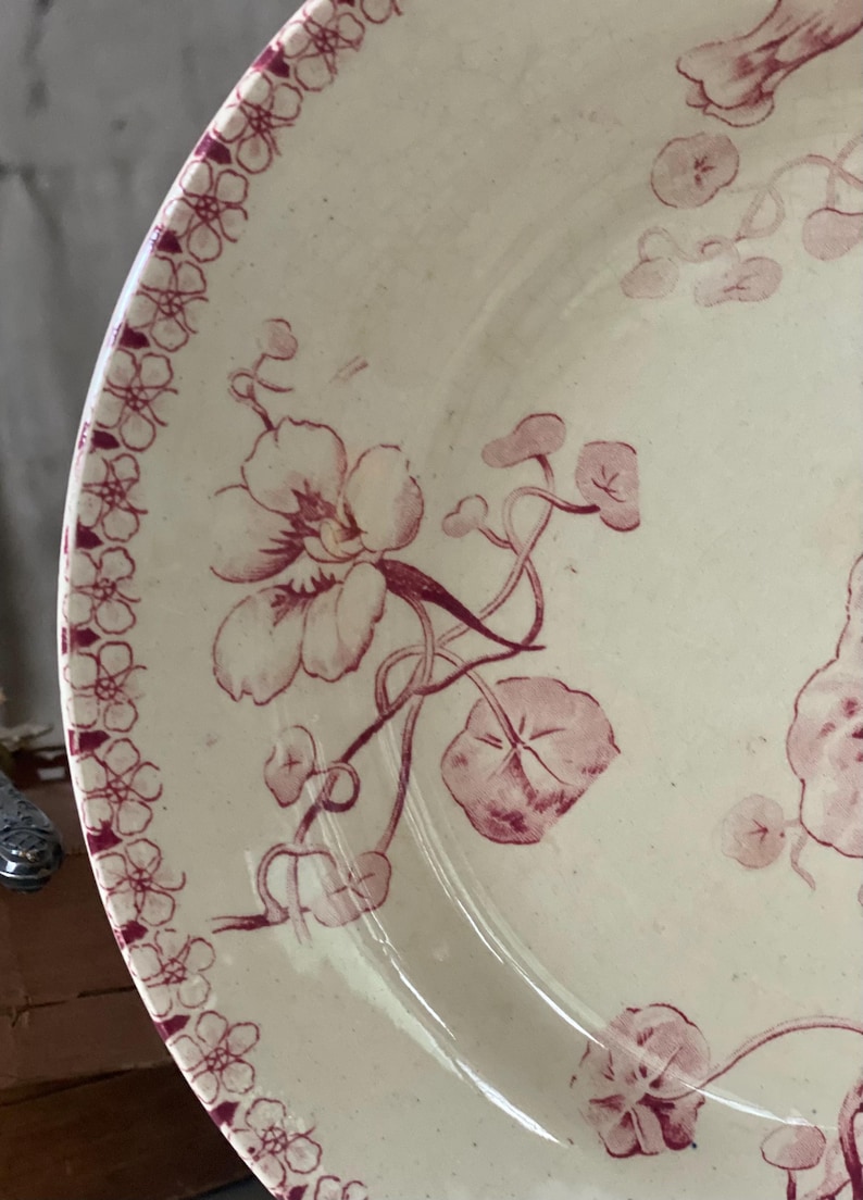 May include: Cream-colored ceramic plate with a floral design in burgundy. The plate features a border of small floral patterns and larger flowers with vines and leaves. A classic, vintage aesthetic.