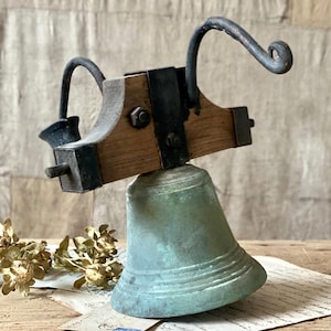 Puede incluir: Campana de bronce antigua con yugo de madera y detalles en metal negro. La campana tiene un aspecto envejecido y desgastado, con un asa curvada y un pequeño badajo. El yugo de madera está asegurado con correas y remaches metálicos.