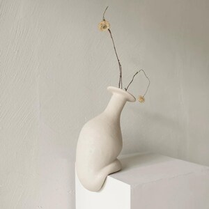 Peut inclure: Un vase en céramique blanc cassé mat avec une forme organique asymétrique qui se courbe sur le bord d'une surface. Le vase présente un col étroit et contient deux tiges brunes fines avec de petites fleurs séchées de couleur claire au sommet.