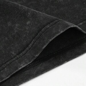 Puede incluir: Primer plano de una camiseta negra con lavado &aacute;cido. La tela tiene un aspecto texturizado con &aacute;reas m&aacute;s claras, creando un efecto descolorido. Se ve el cuello, con una costura de doble pespunte. La camiseta est&aacute; hecha de un material suave y c&oacute;modo.