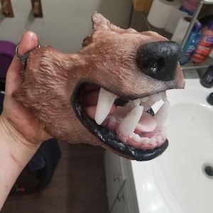 Può includere: Una maschera da lupo marrone e nera con grandi denti bianchi e un naso nero. La maschera ha una superficie strutturata ed è tenuta da un anello di metallo.