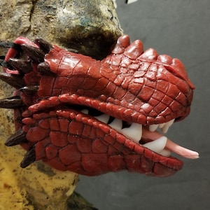 May include: A red dragon mask with detailed scales, sharp teeth, and a pink tongue. The mask has dark brown spikes along the top and sides, and is designed to be worn.