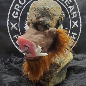 May include: A detailed, grotesque mask of a boar's head. The mask features a pink snout with a silver ring, white tusks, and a brown face with orange fur. The head is painted with mottled colors and has a black strap.