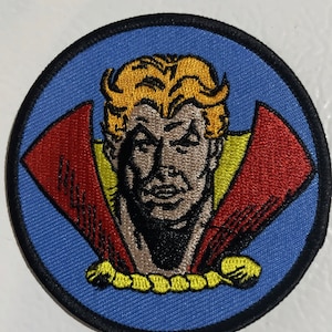 Op de afbeelding: Ronde geborduurde patch met een man met een streng gezicht, blond haar en een rode cape op een blauwe achtergrond. De patch heeft een zwarte rand en gele accenten, die doet denken aan een vintage stripboek.