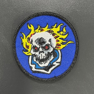 May include: Round embroidered patch featuring a skull with flames and red eyes. The skull is white with black shading, set against a blue background with a black border. The flames are yellow and orange.