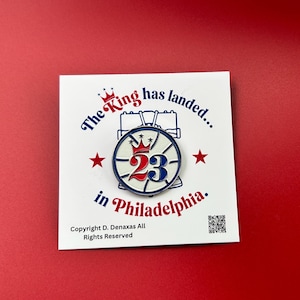 Puede incluir: Un pin esmaltado circular con un gráfico de baloncesto, una corona roja y el número 23 en rojo y azul. Está montado sobre una tarjeta blanca con el texto The King has landed in Philadelphia, un código QR pequeño e información de copyright.