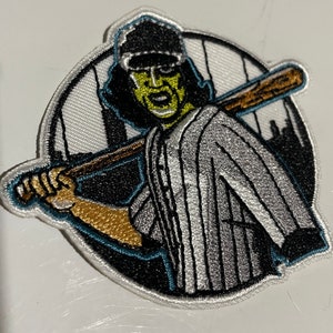Puede incluir: Parche bordado con un jugador de béisbol con cara verde, pelo negro y uniforme a rayas. El parche tiene un fondo blanco y negro con una silueta de paisaje urbano y un borde turquesa.