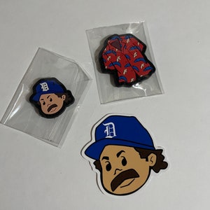 Puede incluir: Tres dijes decorativos. Uno muestra un personaje de dibujos animados con bigote y una gorra de béisbol azul con una "D" blanca. Otro es una camisa hawaiana roja con detalles azules. Todos los dijes están en bolsas de plástico transparente.