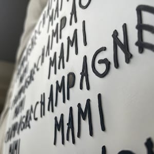 May include: White rectangular sign with the words "CHAMPAGNE MAMI" in a repeating pattern. The text is in a bold, black, handwritten-style font. The sign is angled, showing a side view.