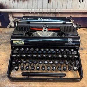 Op de afbeelding: Een zwarte Olympia Progress typemachine met een QWERTY-toetsenbordindeling. Het apparaat heeft een metalen frame, ronde zwarte toetsen met witte letters, een papierwagen en een klein Berretty Nijmegen-label aan de linker voorzijde.