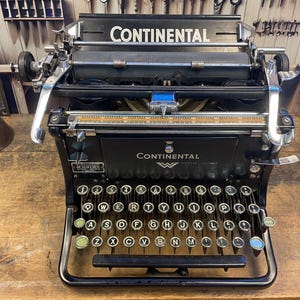 May include: A vintage black Continental typewriter with silver accents and a full QWERTY keyboard. The word "CONTINENTAL" is displayed prominently on the front. The typewriter sits on a wooden surface, showcasing its classic design.