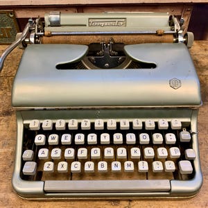 May include: A vintage green Torpedo typewriter with a white keyboard and black keys. The typewriter has a silver return lever and a silver carriage return lever.