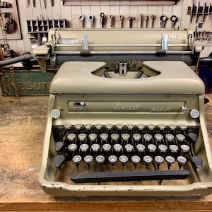 May include: A vintage grey typewriter with black keys and a lever on the left side. The typewriter is labelled "Everest" and "mod. 5T".