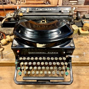 Puede incluir: Una máquina de escribir vintage negra Continental Silenta con un teclado verde y dorado. La máquina de escribir está sobre una mesa de madera.