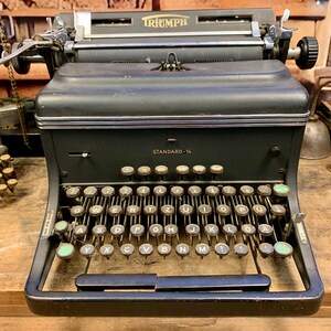 Può includere: Una macchina da scrivere Triumph Standard-14 nera con una tastiera verde e nera. La macchina da scrivere ha un corpo in metallo e una targhetta argentata.