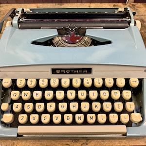 May include: A light blue Brother typewriter with a white keyboard and black keys. The typewriter has a carriage return lever on the left side and a paper release lever on the right side. The typewriter has a black ribbon and a black metal frame.