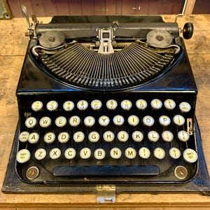 以下が含まれることがあります： 金属製のフレームと白黒のキーボードが付いた黒いビンテージタイプライター。キーには黄色の文字が書かれています。タイプライターにはキャリッジリターンレバーとスペースバーがあります。
