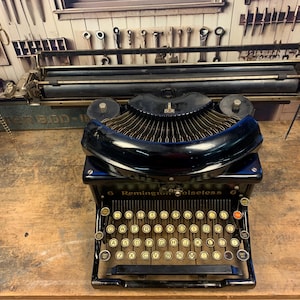 May include: A black vintage Remington Noiseless 6 typewriter with a black metal frame and a yellowed ivory keyboard. The typewriter is sitting on a wooden surface.