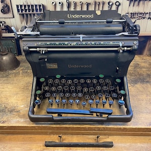 May include: A vintage black Underwood typewriter with a full keyboard and green and blue keys. The word "Underwood" is visible on the front. The typewriter sits on a wooden surface.