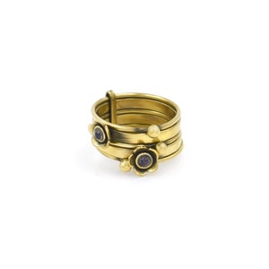 Pode incluir: Um anel dourado com um design de várias faixas. O anel apresenta pequenos detalhes florais com centros roxos e detalhes esféricos. O anel está sobre um fundo branco.
